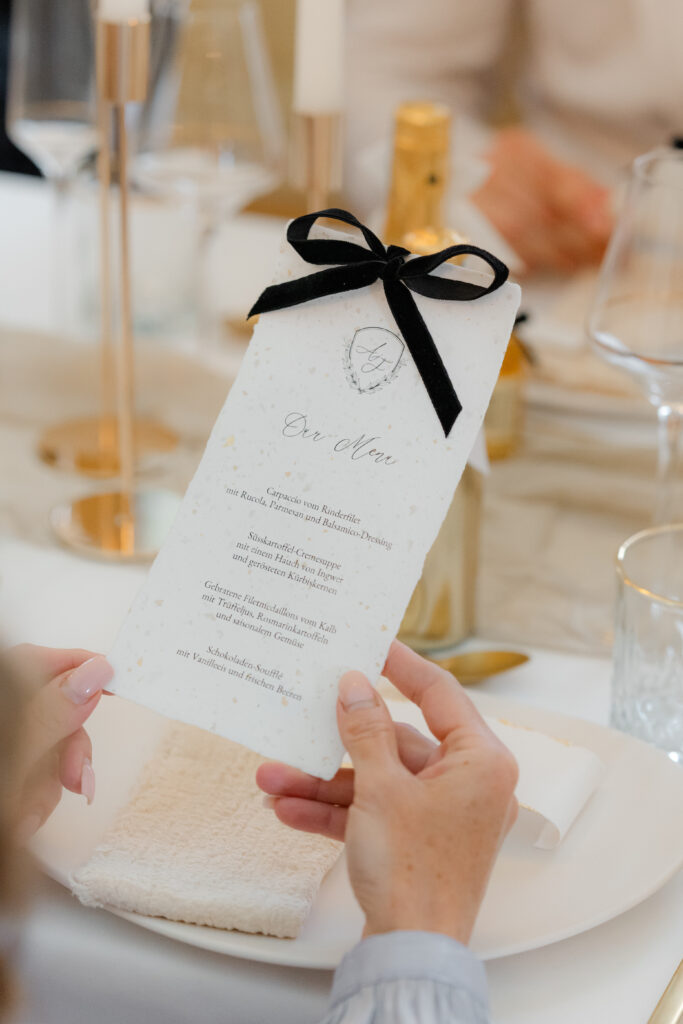 Menükarte auf handgeschöpftem Papier mit eingearbeitetem Blattgold mit Hochzeitswappen und schwarzer Samtschleife passend zum Thema Old Money